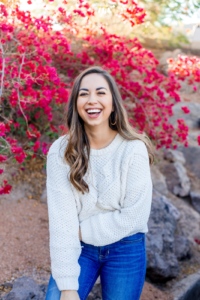 woman with long brown hair laughing in front of a pink flowering bush